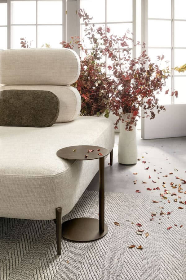 divano-avalon-collezione-divine-7-600x900 Elegante divano color panna con cuscini decorativi scuri, accanto a una grande pianta con foglie rosse in un ambiente luminoso, ideale per arredare con stile e comfort.