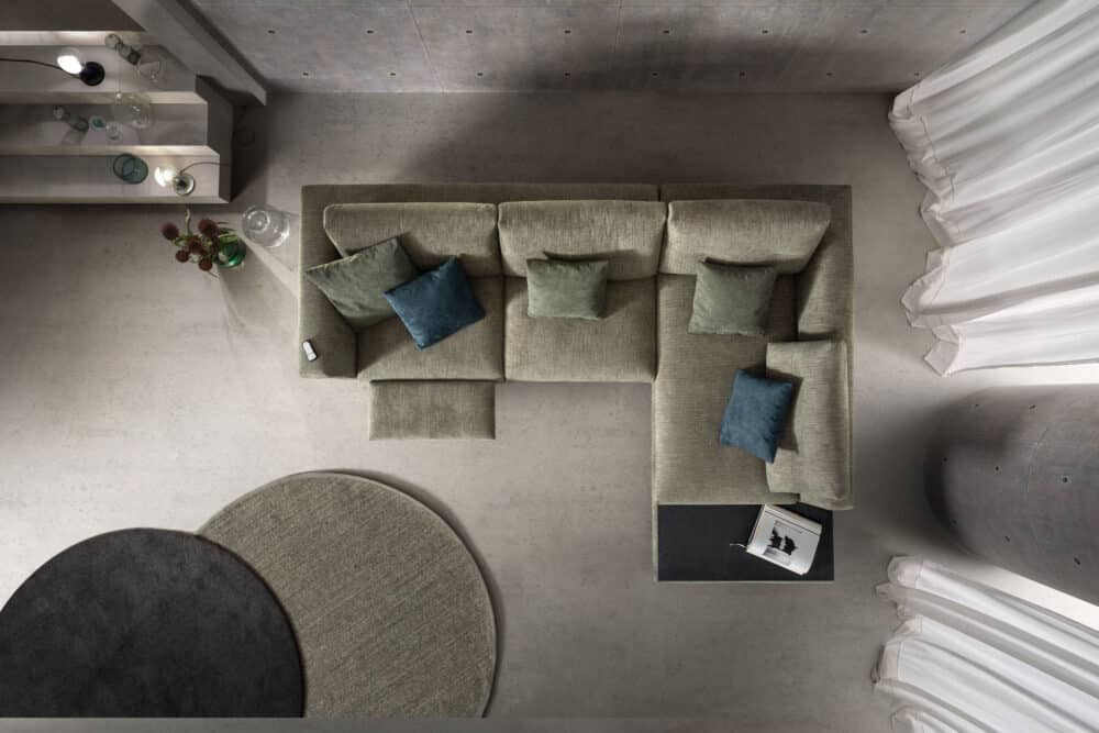 divano-dafne-collezione-experience-2-1000x667 Soggiorno moderno con divano angolare in tessuto beige, cuscini blu e verdi, in un ambiente minimalista con pareti di cemento e tende bianche, arredamento di alta qualità.