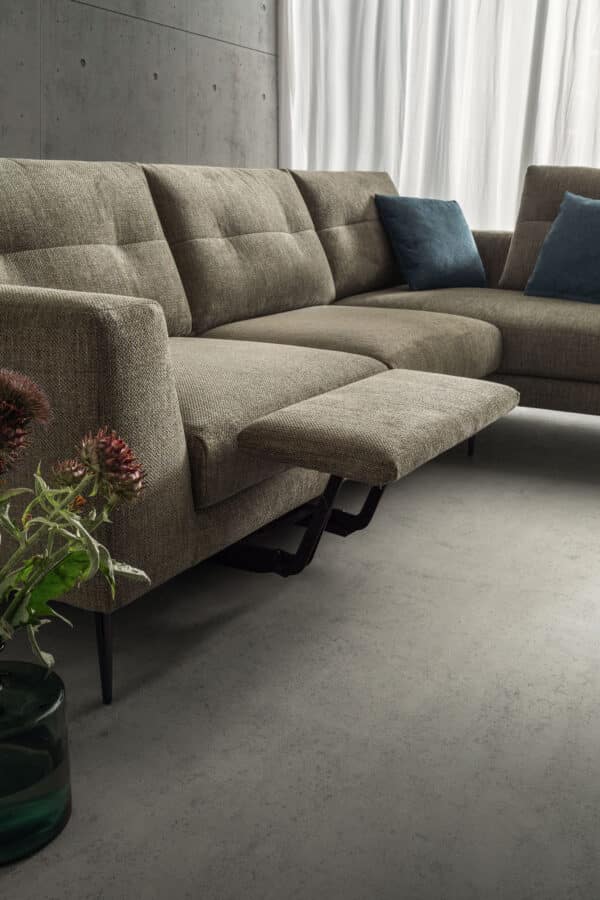 divano-dafne-collezione-experience-3-600x900 Seduta moderna in tessuto beige con poggiapiedi estraibile, abbinata a cuscini blu, in un ambiente minimalista con pareti in cemento e tende bianche.