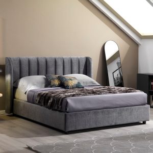 Letto matrimoniale con testiera imbottita grigia, decorato con cuscini e copriletto in pelliccia, in una camera moderna con specchio a terra e pareti beige.