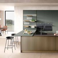 Cucina moderna con ante verde scuro, isola centrale con piano in pietra e sgabelli neri, scaffali aperti con bicchieri e piante, grande finestra con vista sul giardino.