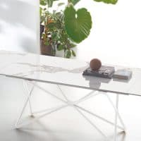 Tavolo moderno in marmo bianco con struttura in metallo bianco, ambientato in uno spazio luminoso con piante verdi sullo sfondo.
