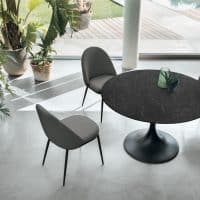 Tavolo rotondo nero con quattro sedie grigie in un ambiente moderno con grandi finestre e piante verdi.