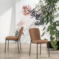 Due sedie AKITA in tessuto beige con struttura in metallo nero, posizionate in un ambiente luminoso con piante verdi e decorazioni astratte alle pareti.