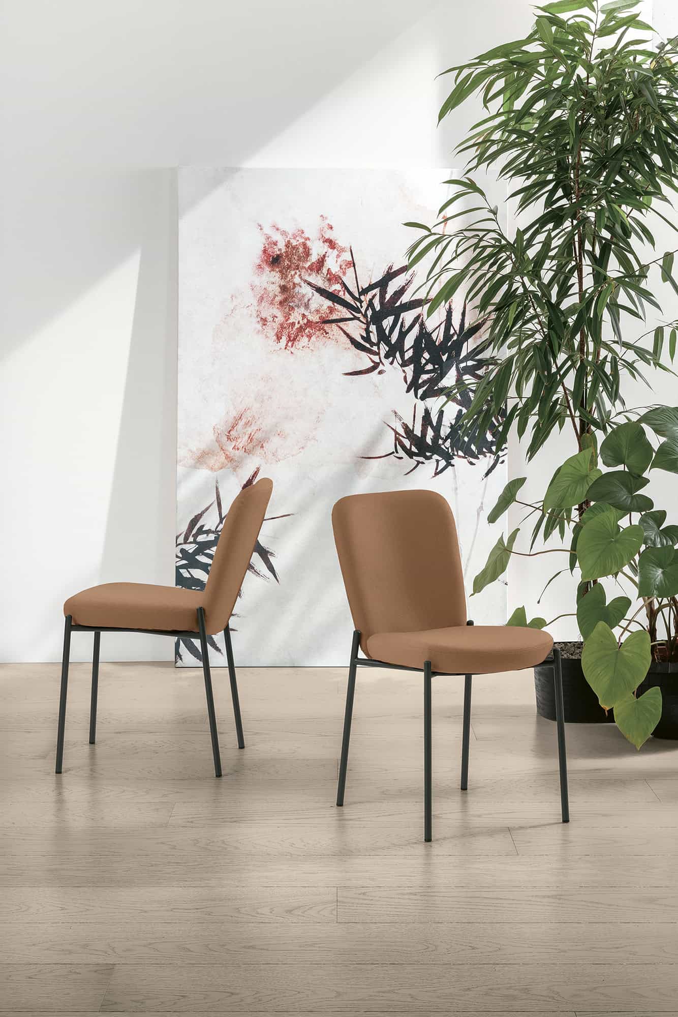 Due sedie AKITA in tessuto beige con struttura in metallo nero, posizionate in un ambiente luminoso con piante verdi e decorazioni astratte alle pareti.